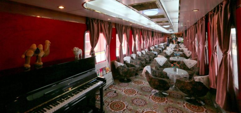 Un luxueux salon du Train Shangri La Express avec une moquette à motifs, des tables rondes en verre, des fauteuils, des rideaux rouges, un piano noir et des figurines de chameaux sur le dessus. Un éclairage chaleureux et des fenêtres bordent le côté droit du wagon.