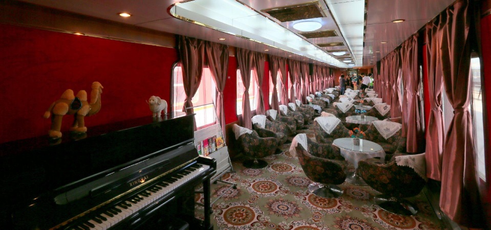 Un luxueux salon du Train Shangri La Express avec une moquette à motifs, des tables rondes en verre, des fauteuils, des rideaux rouges, un piano noir et des figurines de chameaux sur le dessus. Un éclairage chaleureux et des fenêtres bordent le côté droit du wagon.