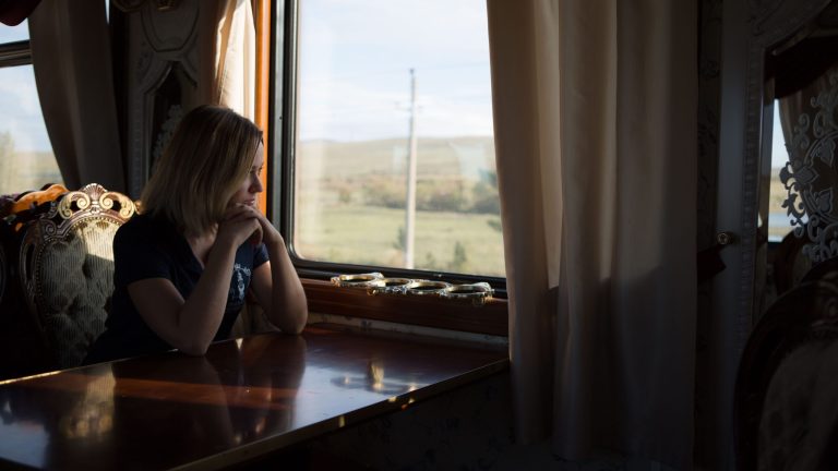 Une femme est assise seule à une table en bois près d'une fenêtre à rideaux, regardant pensivement à l'extérieur tandis que le train Russie Impériale glisse le long de champs verts éclairés par le soleil et de collines lointaines.