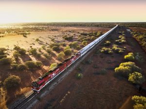 Le Ghan, un long train de passagers aux locomotives rouges, traverse l'outback australien au coucher du soleil, entouré d'arbres clairsemés et d'un paysage de terre rouge saisissant.