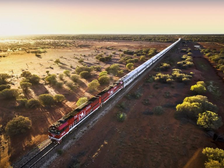 Le Ghan, un long train de passagers aux locomotives rouges, traverse l'outback australien au coucher du soleil, entouré d'arbres clairsemés et d'un paysage de terre rouge saisissant.