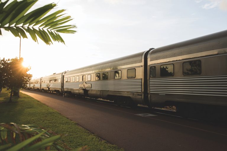 Un train argenté Great Southern circule sur des voies à côté d'une pelouse verte et de palmiers pendant le coucher du soleil, la lumière douce du soleil illuminant la scène.