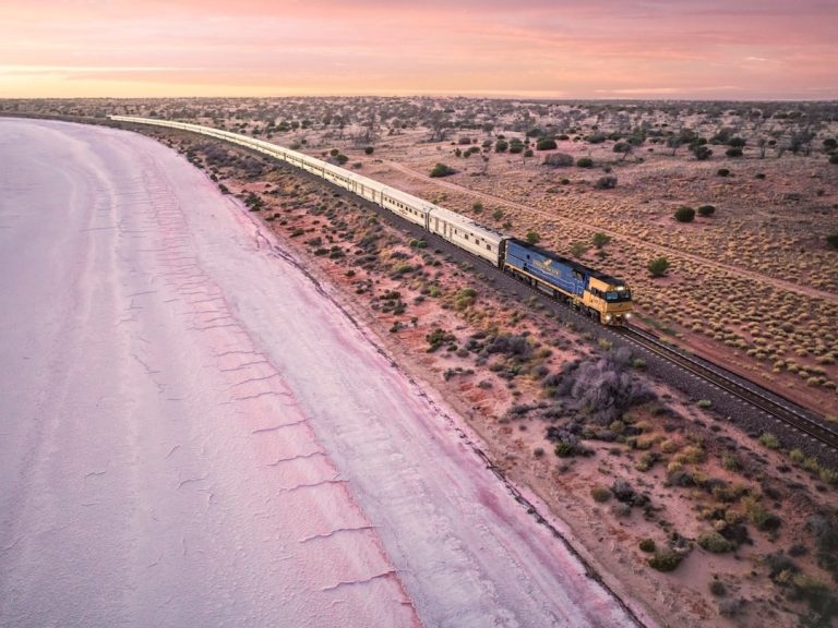 Le train de passagers de l'Indian Pacific glisse sur les voies à côté d'un lac salé de couleur rose et d'un paysage aride sous un ciel de coucher de soleil pastel. Une végétation clairsemée parsème la terre, s'étirant vers l'horizon.