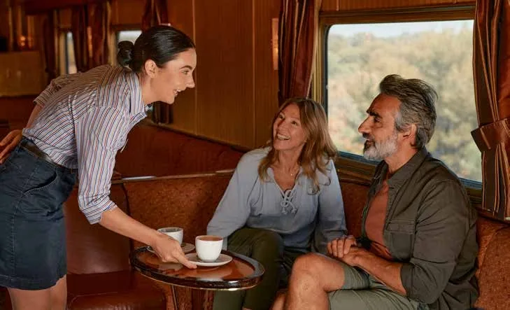 Hôtesse servant un couple dans le salon d'un train de luxe australien Journey Beyond avec vue sur la forêt